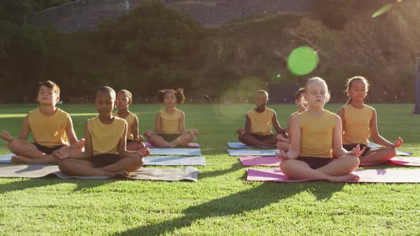 Diverse group of schoolchildren sitting on mats meditating during yoga lesson outdoors