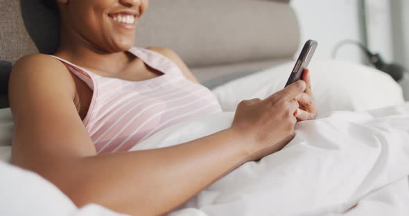 Happy african american woman laying in bed, using smartphone in bedroom