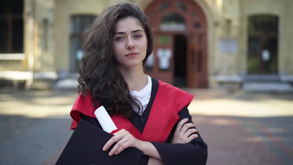 Gorgeous Confident Caucasian Woman in Graduation Toga Looking at Camera Standing with Diploma