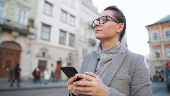 Woman with Glasses Wearing a Coat Walking Down an Old Street and Using Smartphone