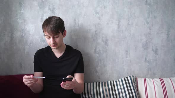 Young Man Checks the Information in Phone About the Measured Temperature. Checking Symptoms