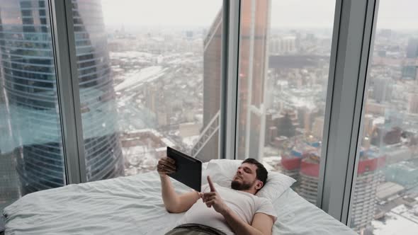 Sleepy Man Lying on the Bed with a Tablet in His Hands in a Modern Apartment Near the Window