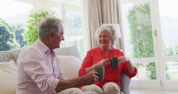 Happy caucasian senior couple sitting in sunny living room talking and drinking cups of coffee