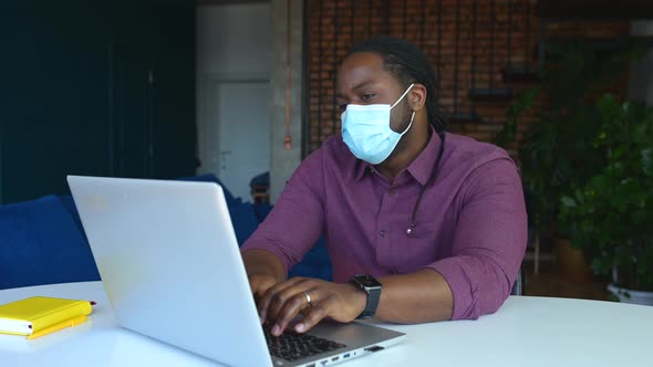 Concentrated AfricanAmerican Male Employee Wearing Medical Mask Using Laptop