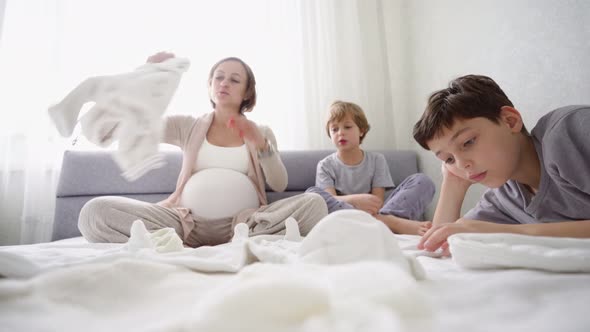 Pregnant Woman Sits on the Bed with Two Sons and Prepares Things for the Birth of a Child