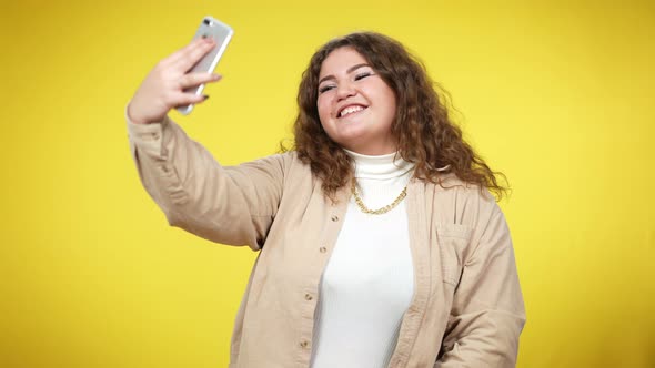 Cheerful Plussize Millennial Woman Taking Selfie on Smartphone at Yellow Background