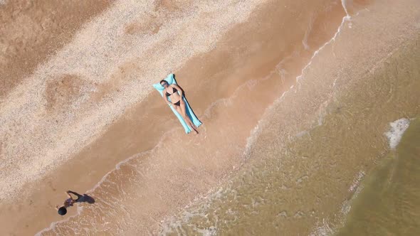 Woman in a Black Swimsuit Lies on a Blue Mattress on a Sandy Beach By the Sea