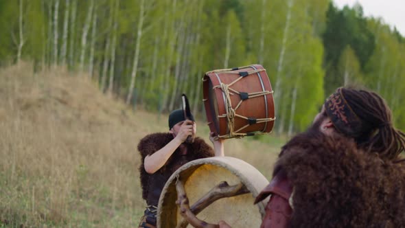 Man in Folk Clothes Taps Drum with Friend Against Forest