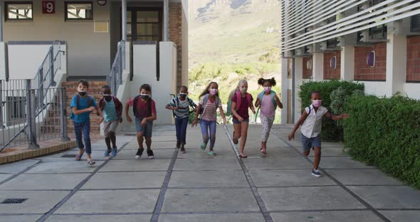 Diverse group of schoolchildren wearing backpacks and face masks running at school yard