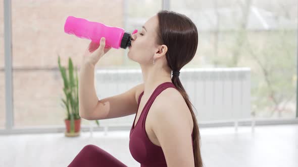 a Young Woman in a Burgundy Tracksuit Drinks Water on a Mat in a Gym