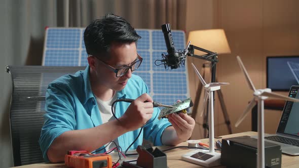 Asian Man With Wind Turbine Fix The Circuit Board While Working With Laptop Next To The Solar Cell