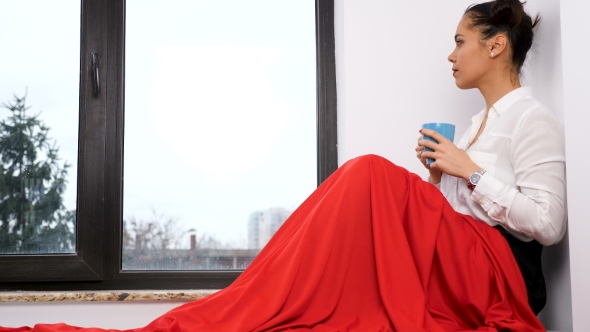 Woman Covered with Red Blanket Sitting Next To a Big Window Drinking Tea