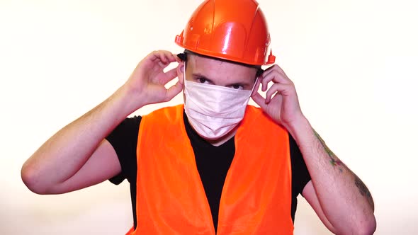 Male Construction Worker in Overalls Putting on Medical Mask on Face on White Background