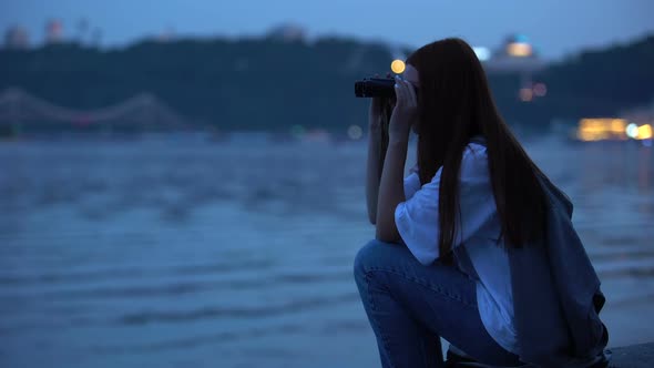 Girl Looking Through Binoculars at Lights of Big City and Ships on Sea, Evening