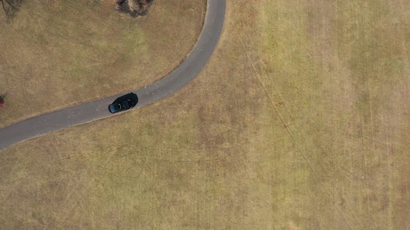 An aerial, top down drone view over a single car driving through an empty park on a sunny day. The d