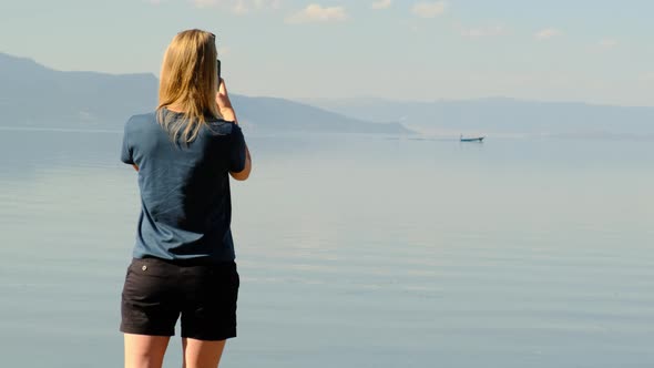 Unrecognizable Blonde Woman in Blue Shirt and Black Short Shorts Using Her Phone Taking Pictures of