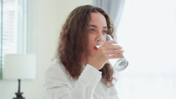 Portrait of Latino woman sit on bed drink a glass of water in bedroom.