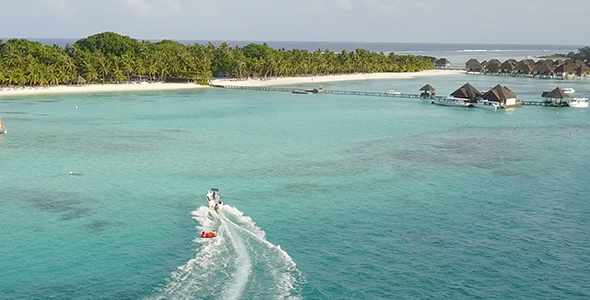 Drone Following Speedboat Towing a Water Donut with People Towards Tropical Island at High Speed