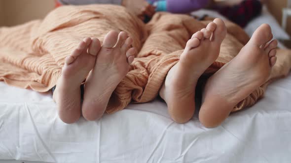 Married couple feet are moving under blanket in bed in funny rhythm, couple is having fun at bedroom