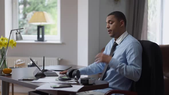 Side View of Overworked Young African American Man Taking Off Headphones Loosing Necktie Sighing