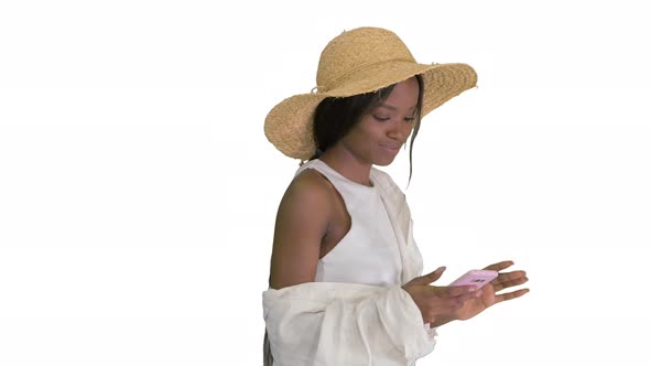 Pretty African American Woman in a Straw Hat Texting on Her Phone While Walking on White Background