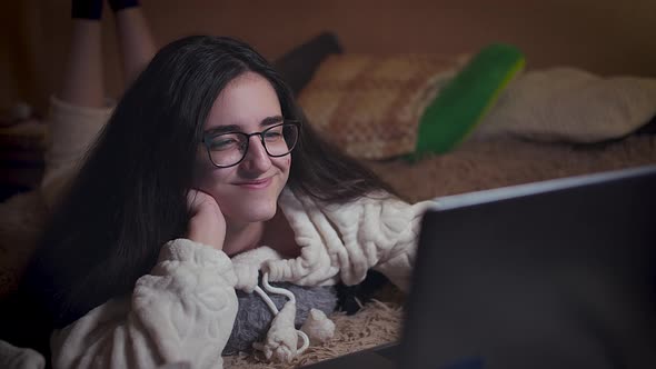 Girl with glasses at night reads the news in a laptop lies in bed