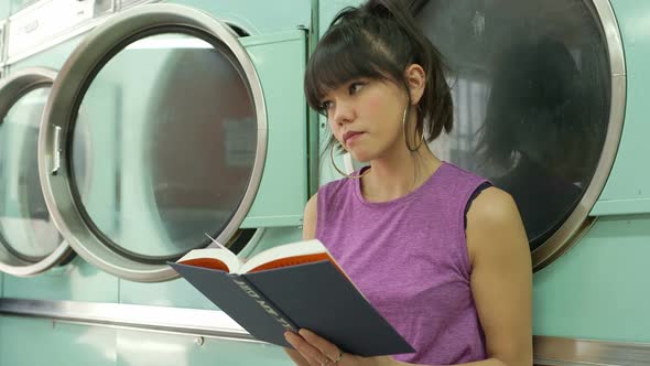 A Young Woman sits reading a book whilst waiting for her laundry