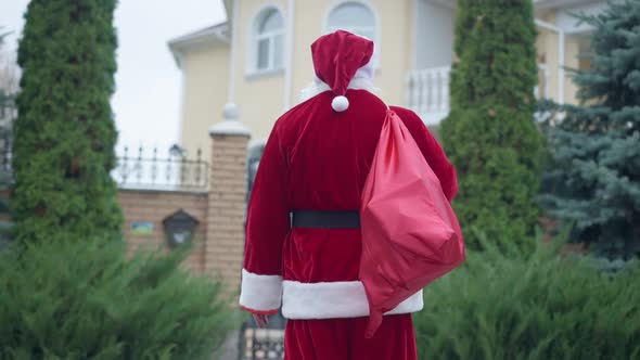 Back View Santa in Red Costume with Gift Bag Standing Outdoors in Front of House Looking Around