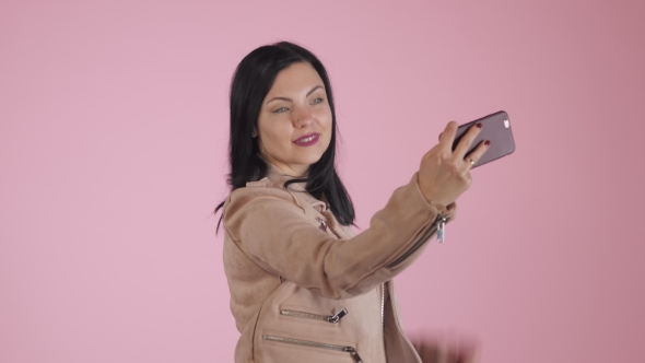 Smiling Happy Brunette Woman in Pink Jacket Making Selfie on Smartphone Over Colorful Background.