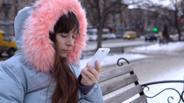 A Young Woman in a Gray Warm Jacket Uses a Phone in the Street in the Winter.
