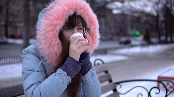 A Young Woman in a Gray Warm Jacket Uses a Phone in the Street in the Winter.