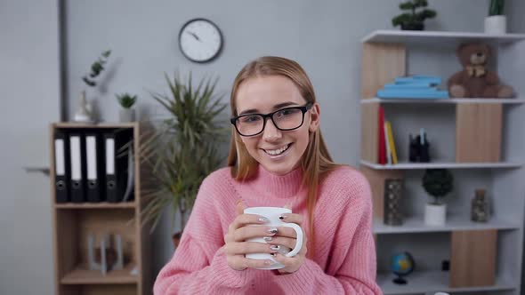 Young Girl in Glasses Dressed in Pink Sweater Holding in Hands Cup of Tea and Looking at Camera