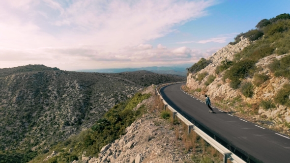 Hipster Man Rides on Skateboard on Mountain Road