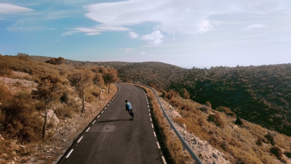 Hipster Man Rides on Skateboard on Mountain Road