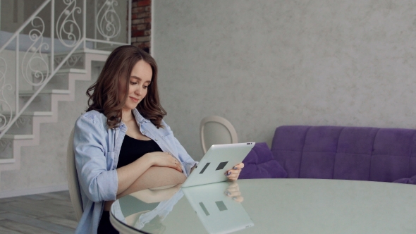 Happy Pregnant Woman Sitting at a Glass Kitchen Table Drinking Coffee and Using a Tablet Computer