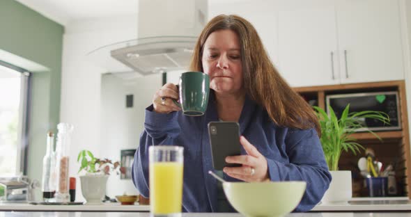 Happy caucasian woman eating breakfast, using smartphone in kitchen