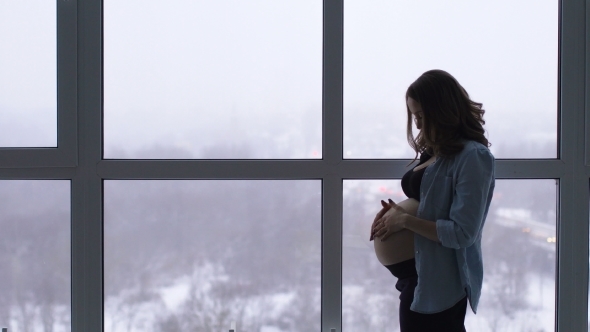 A Happy Pregnant Woman Stands at a Large Panoramic Window Located at a High Altitude He Touches His