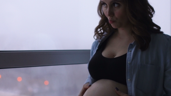 A Happy Pregnant Woman Stands at a Large Panoramic Window Located at a High Altitude. He Touches His