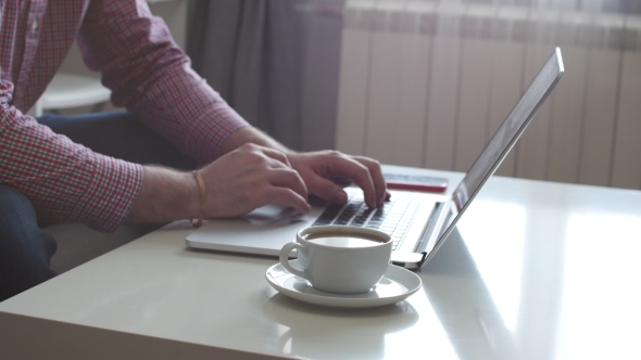 A Person Works From Home Using the Top and Trackpad. Young Business Man Sitting at Table Drinking