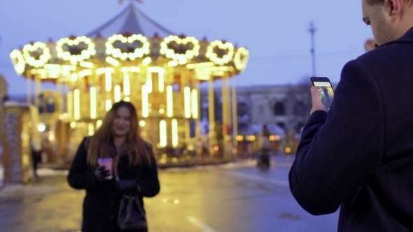 Husband Makes Photo of His Wife Using Phone on the Carousel Background