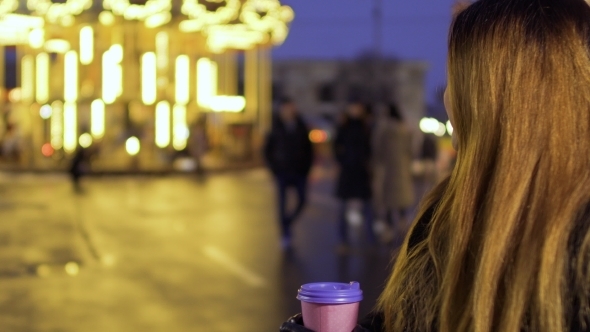 Beautiful Woman with Long Hair Enjoys View of Carousel and Drinks Coffee