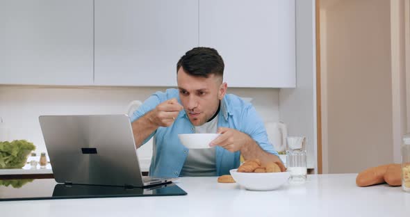 Young Man Having Breakfast in the Cozy Kitchen Simultaneously Working on Computer