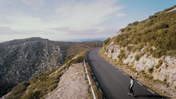 Hipster Man Rides on Skateboard on Mountain Road