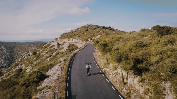 Hipster Man Rides on Skateboard on Mountain Road