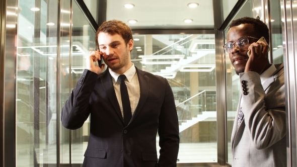 Two Multinational Business Man Standing in Elevator and Use Smartphone. Business People in Large