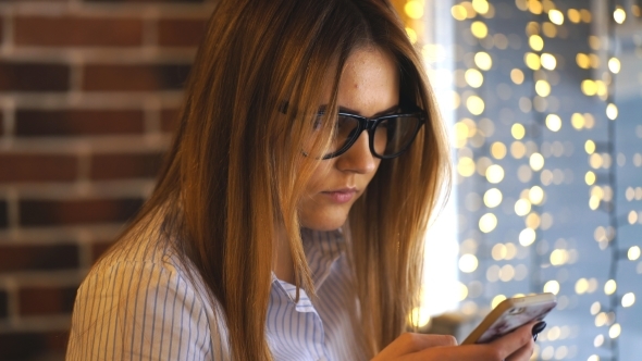 Young Woman Sitting in Coffee Shop at Wooden Table and Using Smartphone