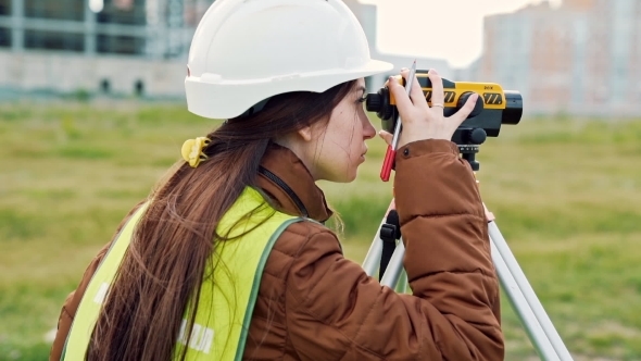 Young Woman Surveyor in Green Work Clothes and Helmet Adjusts the Equipment, Produces Calculations