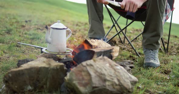 Young Man Sitting in Front of Bonfire. Man Cutting with Knife a Sprig. Coffe on the Fire.