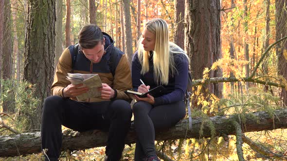 A Hiking Couple Sits on a Broken Tree in a Forest, Man Reads Off a Map and the Woman Writes Notes