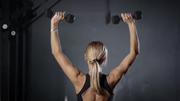 Sporty Young Blonde Woman Is Lifting Dumbbells By Hands in Gym, Standing Back To Camera